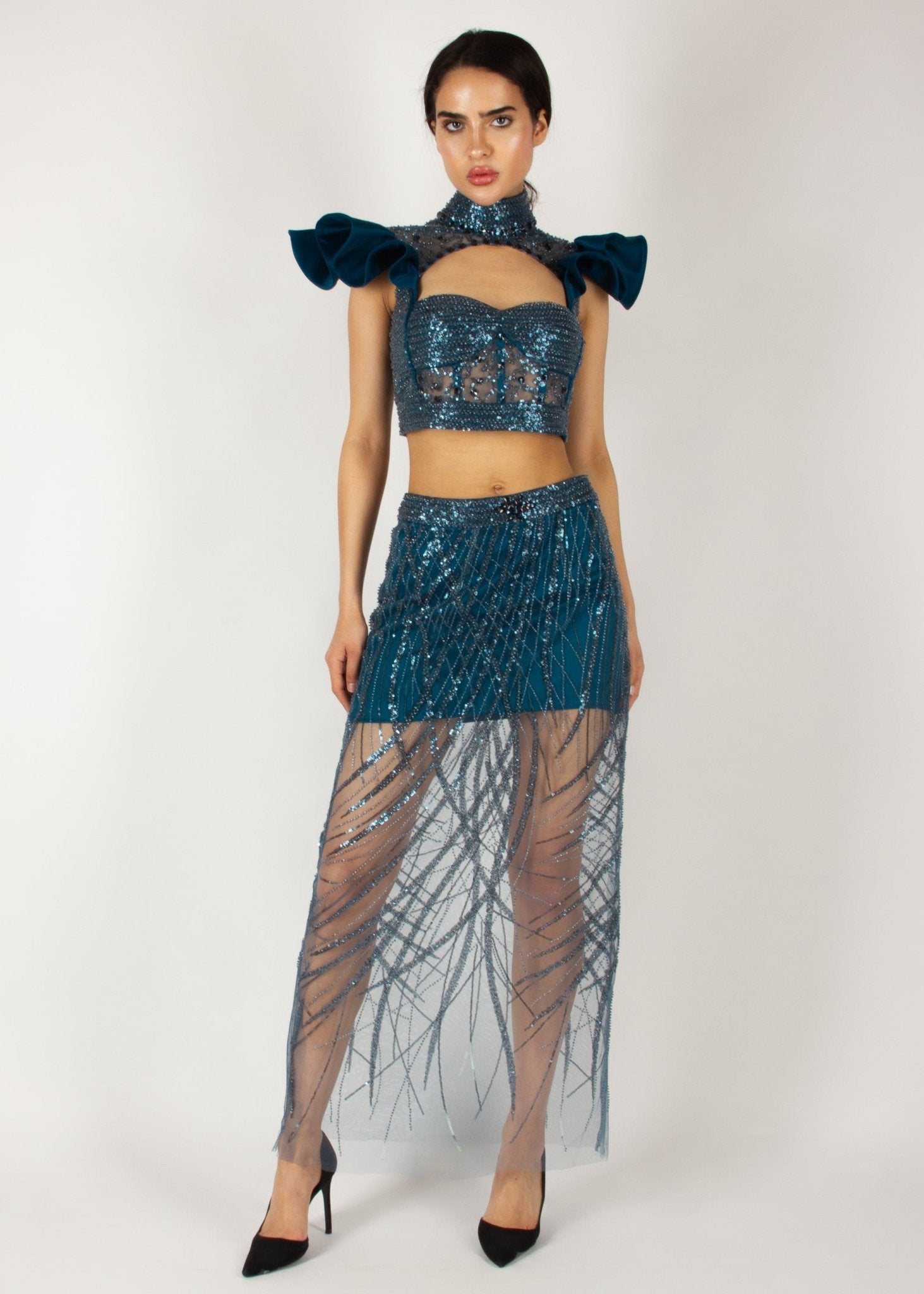 Elegant two-piece couture set by MNour Design, featuring a shimmering blue embellished crop top with dramatic ruffled shoulders and a high-neck cut, paired with a matching sheer beaded skirt for a bold and sophisticated look. Explore this stunning design at www.mnourdesign.com.