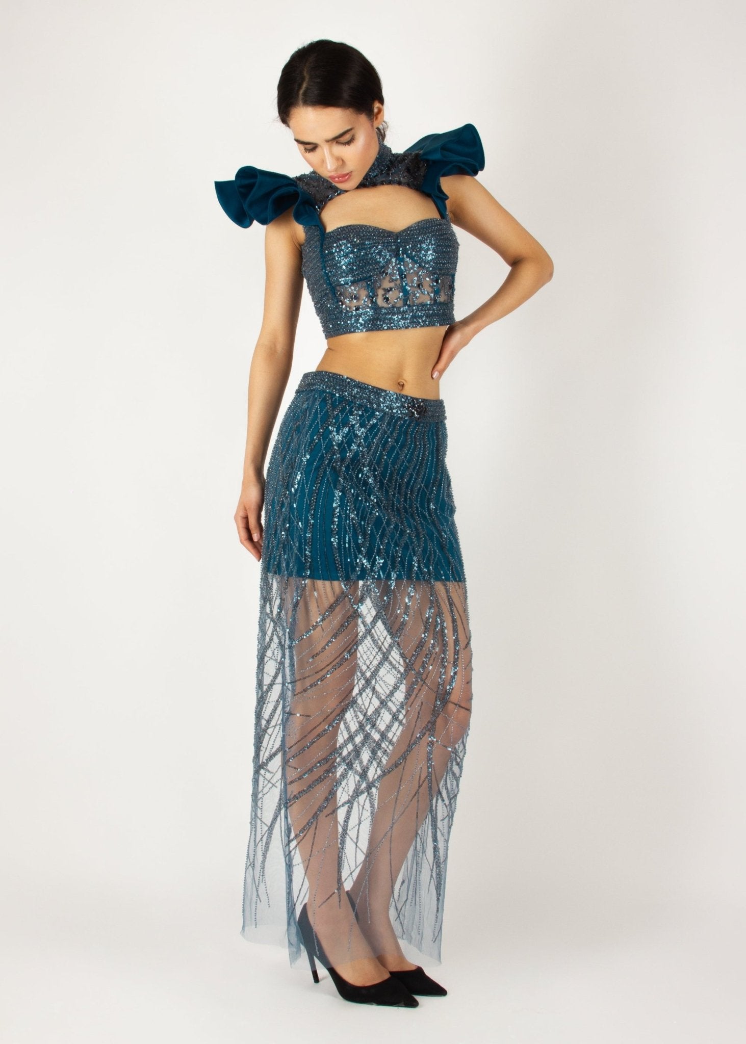 Elegant two-piece couture set by MNour Design, featuring a shimmering blue embellished crop top with dramatic ruffled shoulders and a high-neck cut, paired with a matching sheer beaded skirt for a bold and sophisticated look. Explore this stunning design at www.mnourdesign.com.