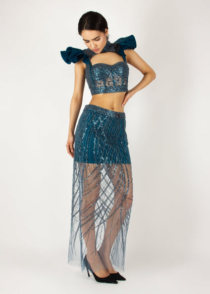 Elegant two-piece couture set by MNour Design, featuring a shimmering blue embellished crop top with dramatic ruffled shoulders and a high-neck cut, paired with a matching sheer beaded skirt for a bold and sophisticated look. Explore this stunning design at www.mnourdesign.com.