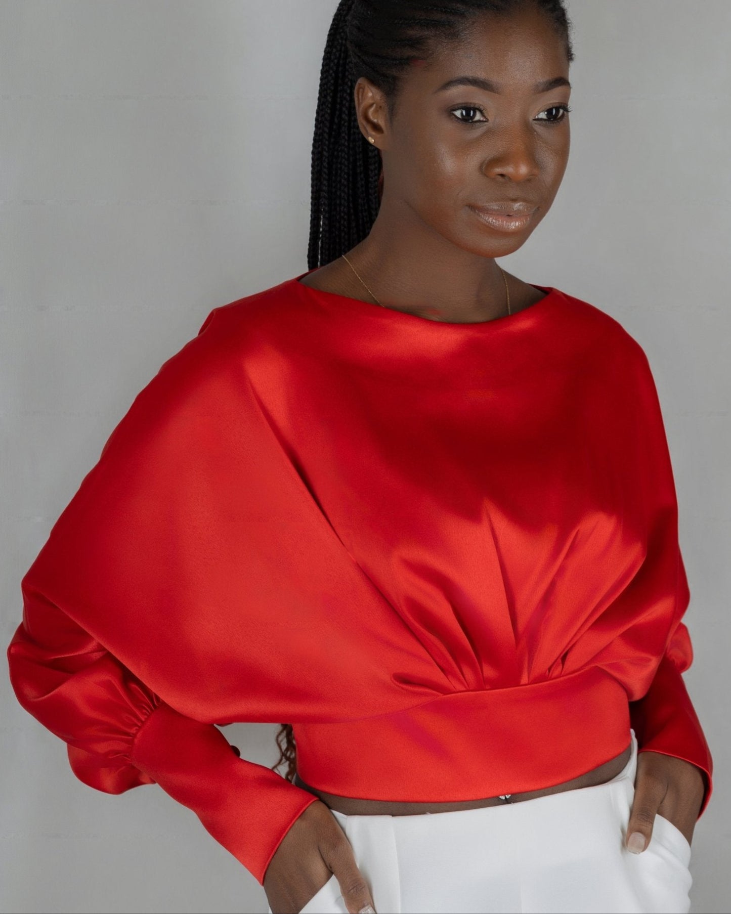 A model wears a bold red, satin-like top from the "HAWAA" collection, specially designed for Valentine's Day. The top features an asymmetrical design with puffed sleeves and a draped neckline that creates a cut-out detail on one side. It is cinched at the waist with a matching belt and paired with black pants. The model stands against a simple gray textured background, emphasizing the vibrant color and elegant silhouette of the outfit.