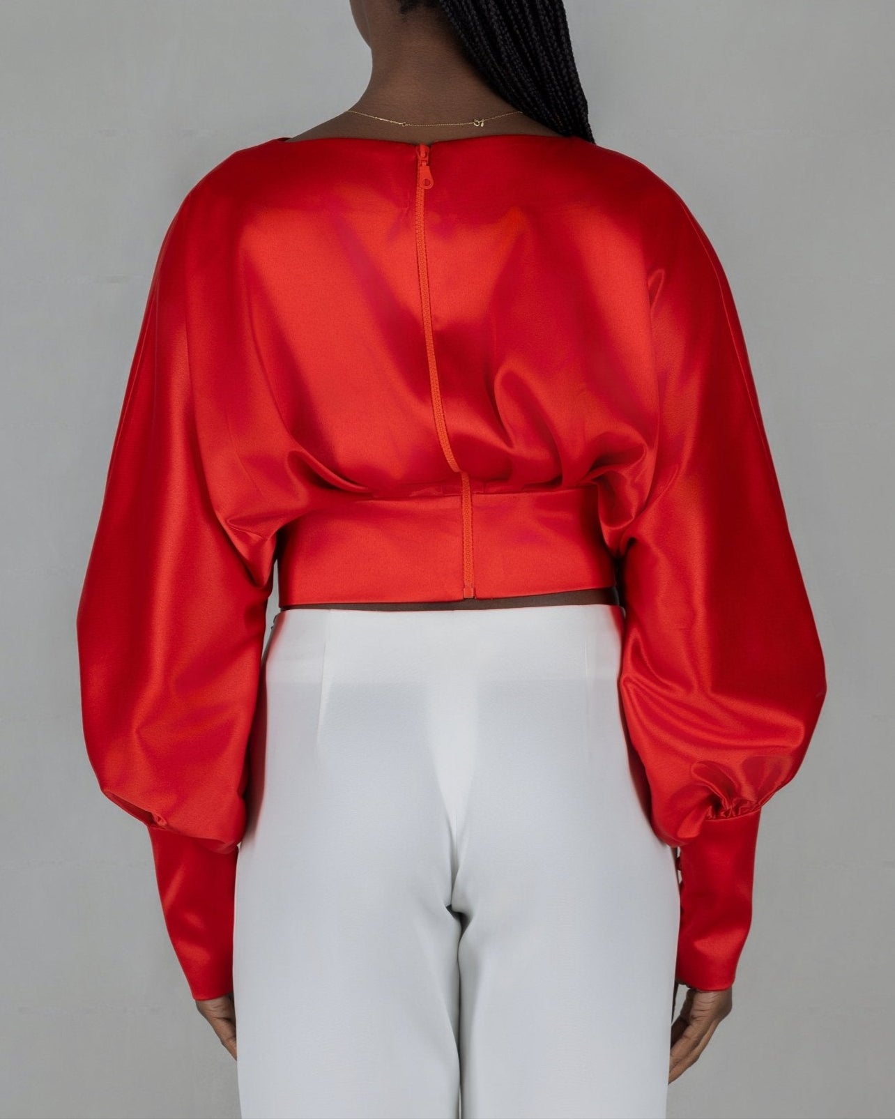 A model wears a bold red, satin-like top from the "HAWAA" collection, specially designed for Valentine's Day. The top features an asymmetrical design with puffed sleeves and a draped neckline that creates a cut-out detail on one side. It is cinched at the waist with a matching belt and paired with black pants. The model stands against a simple gray textured background, emphasizing the vibrant color and elegant silhouette of the outfit.