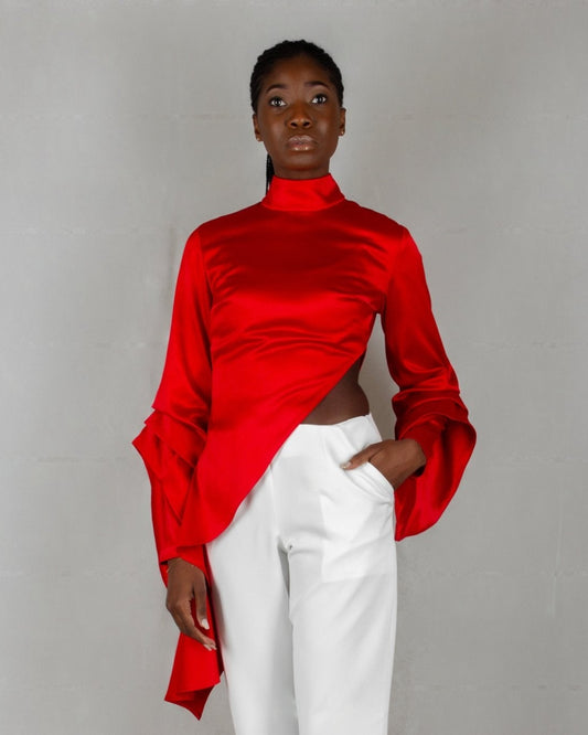 A model showcases a striking outfit from the "HAWAA" collection, specially designed for Valentine's Day. The ensemble features a bold red, high-neck blouse with long sleeves and an asymmetrical design that drapes elegantly over one side. It is paired with sleek white wide-leg pants. The model stands barefoot against a minimalist gray textured background, highlighting the vibrant contrast between the red top and white pants.