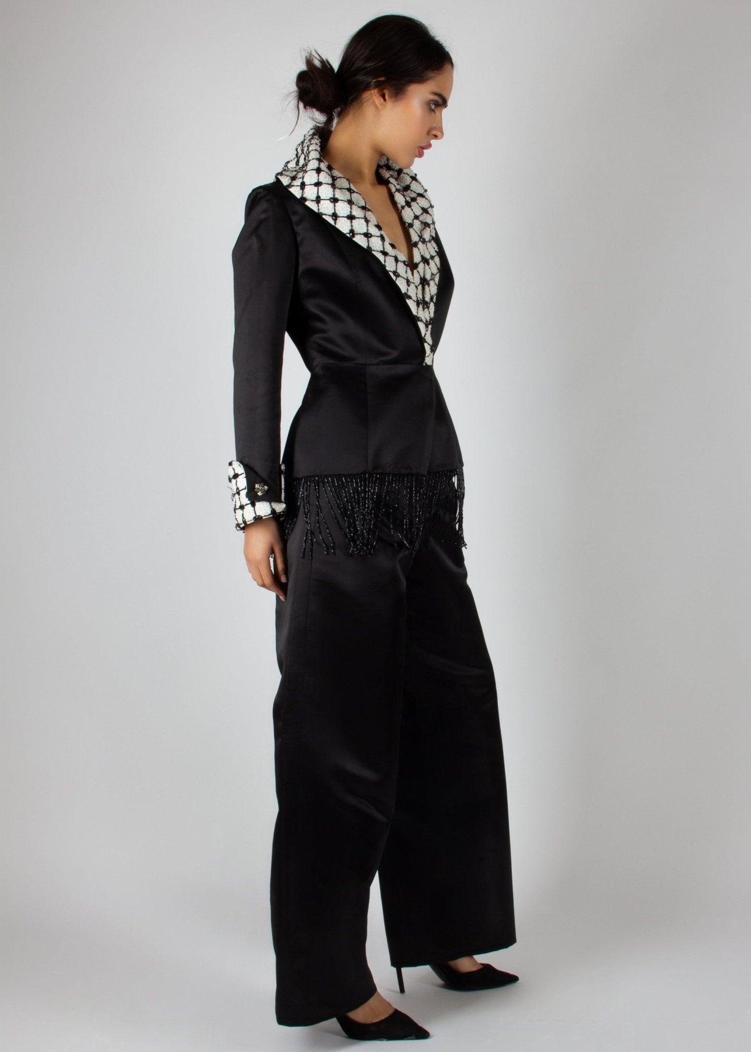 Elegant black satin pantsuit from the HORRA collection by MNour Design, featuring a statement keffiyeh-inspired lapel and cuff details. The tailored blazer is adorned with intricate beaded fringe, adding a touch of modern sophistication. Discover more at www.mnourdesign.com