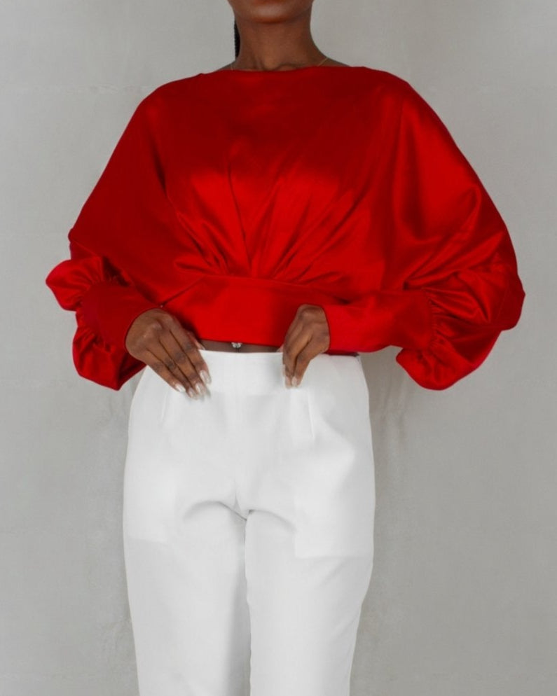 A model wears a bold red, satin-like top from the "HAWAA" collection, specially designed for Valentine's Day. The top features an asymmetrical design with puffed sleeves and a draped neckline that creates a cut-out detail on one side. It is cinched at the waist with a matching belt and paired with black pants. The model stands against a simple gray textured background, emphasizing the vibrant color and elegant silhouette of the outfit.