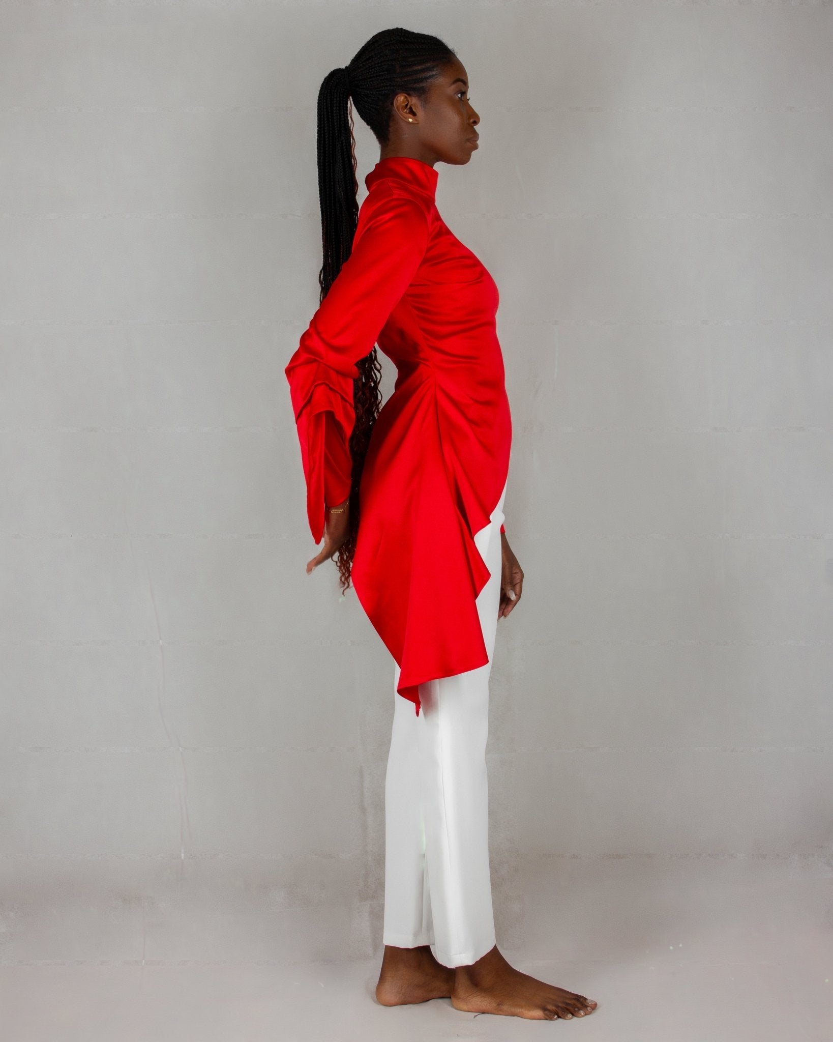 A model showcases a striking outfit from the "HAWAA" collection, specially designed for Valentine's Day. The ensemble features a bold red, high-neck blouse with long sleeves and an asymmetrical design that drapes elegantly over one side. It is paired with sleek white wide-leg pants. The model stands barefoot against a minimalist gray textured background, highlighting the vibrant contrast between the red top and white pants.