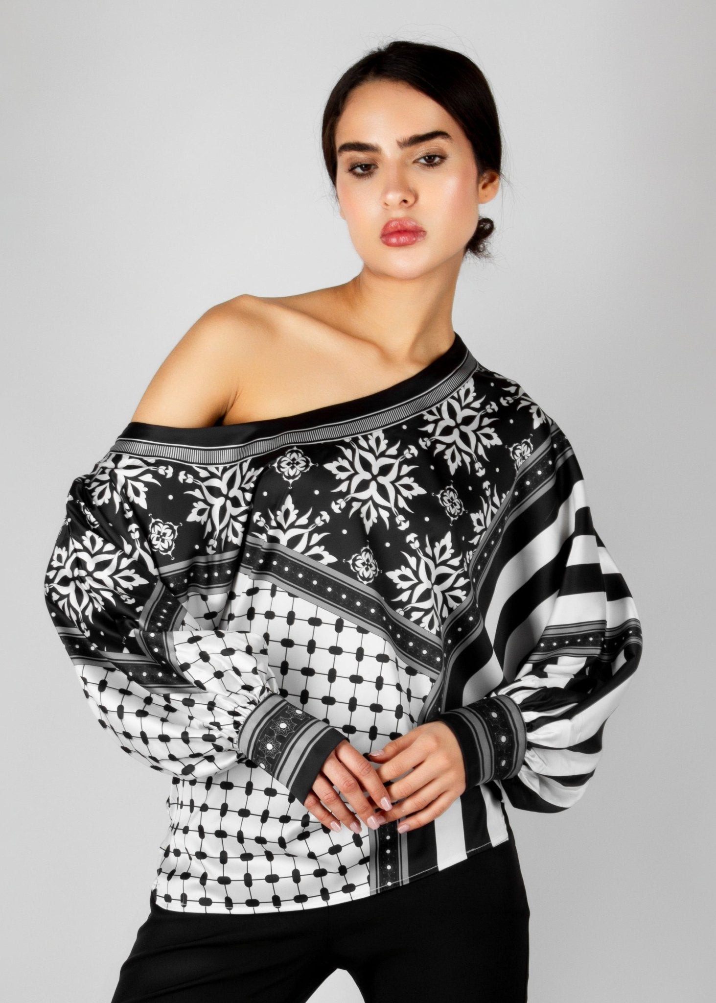 Bold off-shoulder blouse from the HORRA collection by MNour Design, featuring a striking keffiyeh-inspired pattern with intricate geometric details. Paired with sleek black trousers for a modern, sophisticated look. Explore more at www.mnourdesign.com